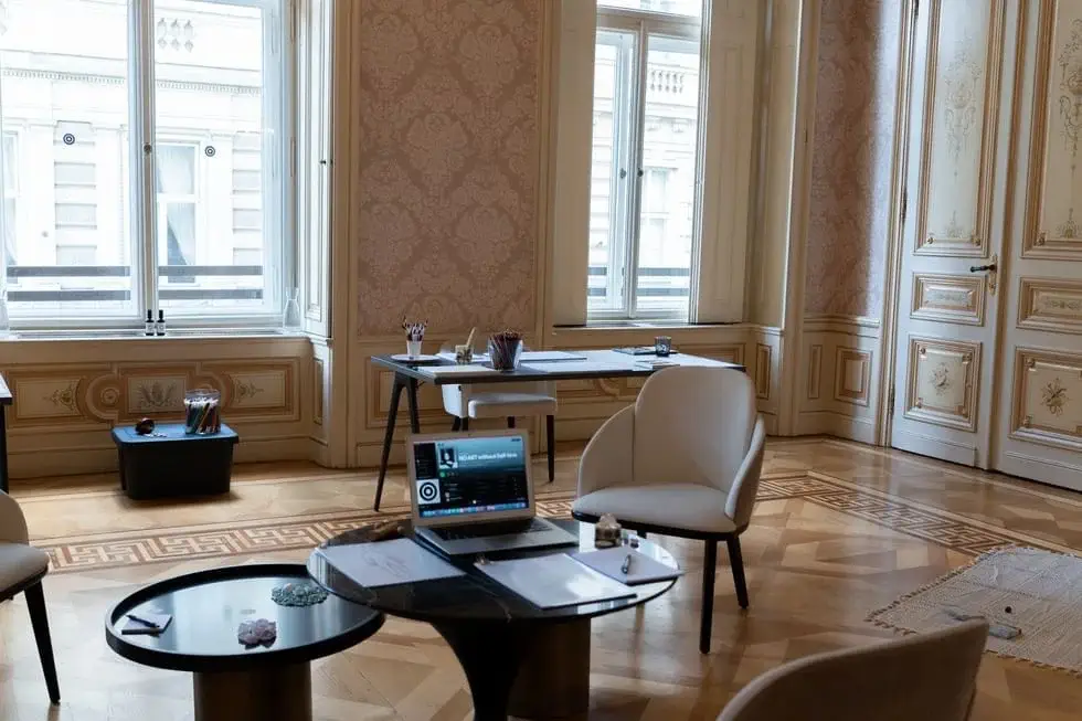 A laptop and papers sit on round tables in an elegant, ornate office with large windows, wooden floors, and classical wall paneling.