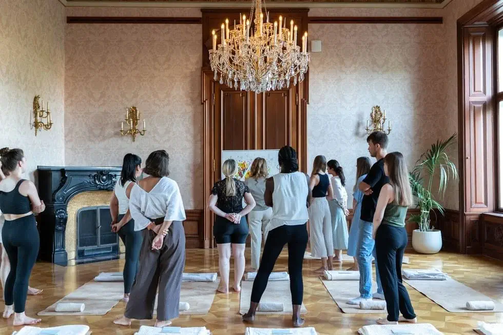 A group of people stand on yoga mats in an elegant room with a chandelier, fireplace, and large windows, facing a person at the front.