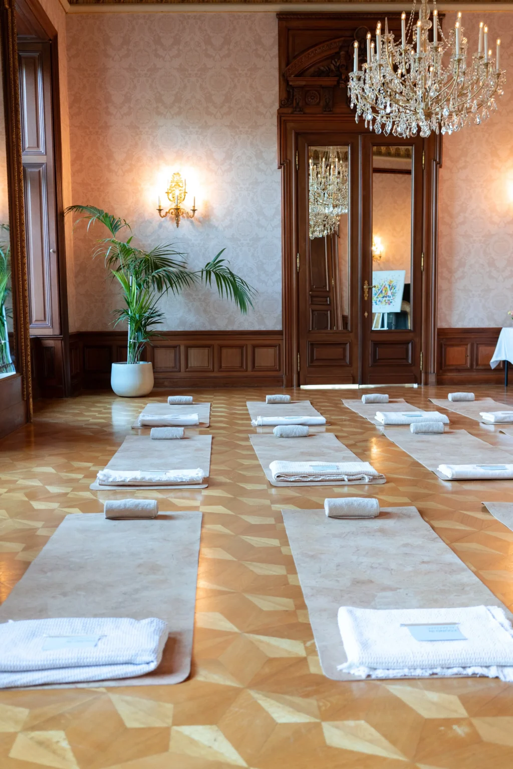 A luxurious room at Almanac Palais, Vienna, with chandeliers, wall sconces, and large mirrors features yoga mats, towels, and bolsters arranged in neat rows on a patterned wooden floor.
