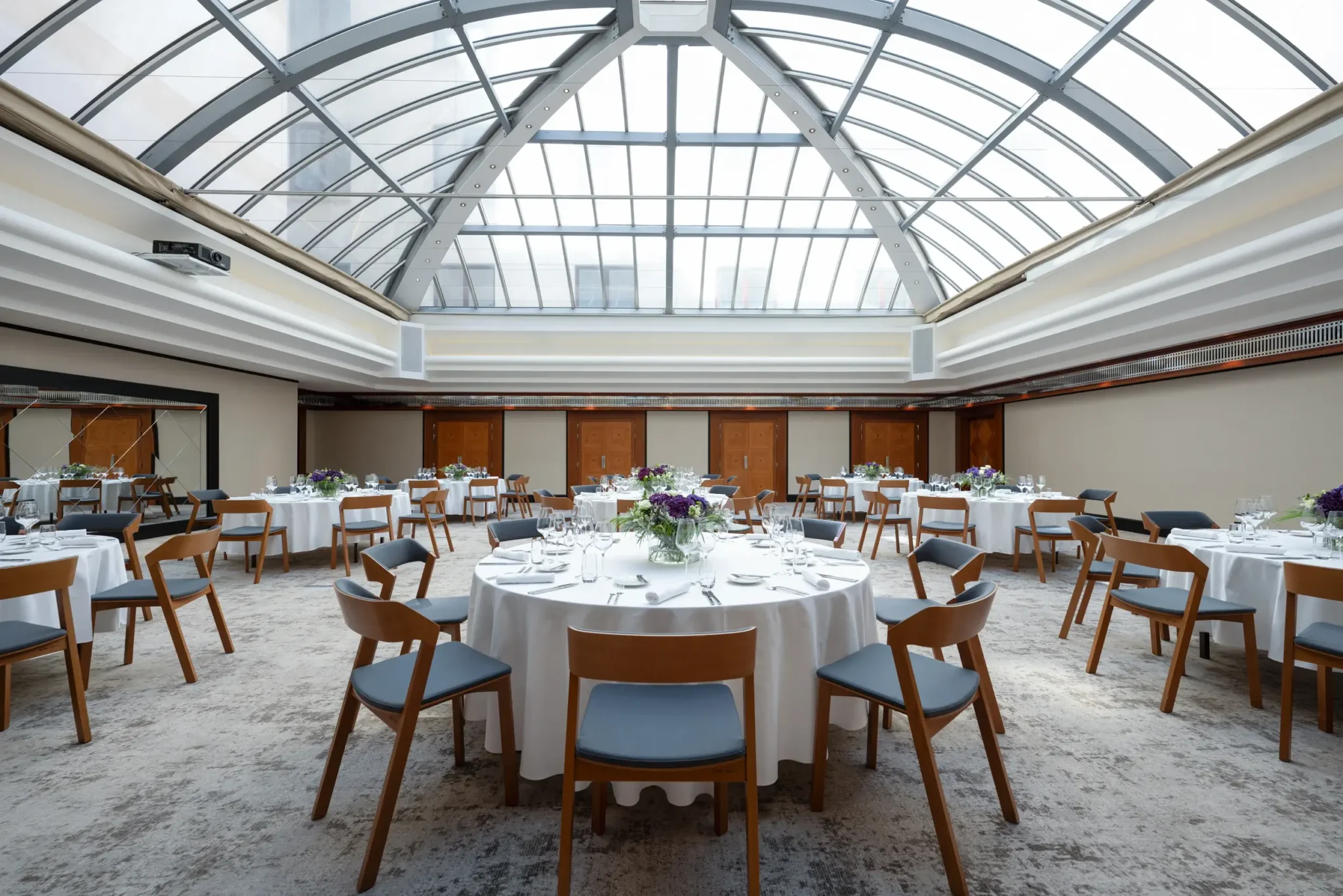 A spacious, well-lit banquet hall with round tables set for dining under a large glass skylight, featuring modern wooden chairs and floral centerpieces.