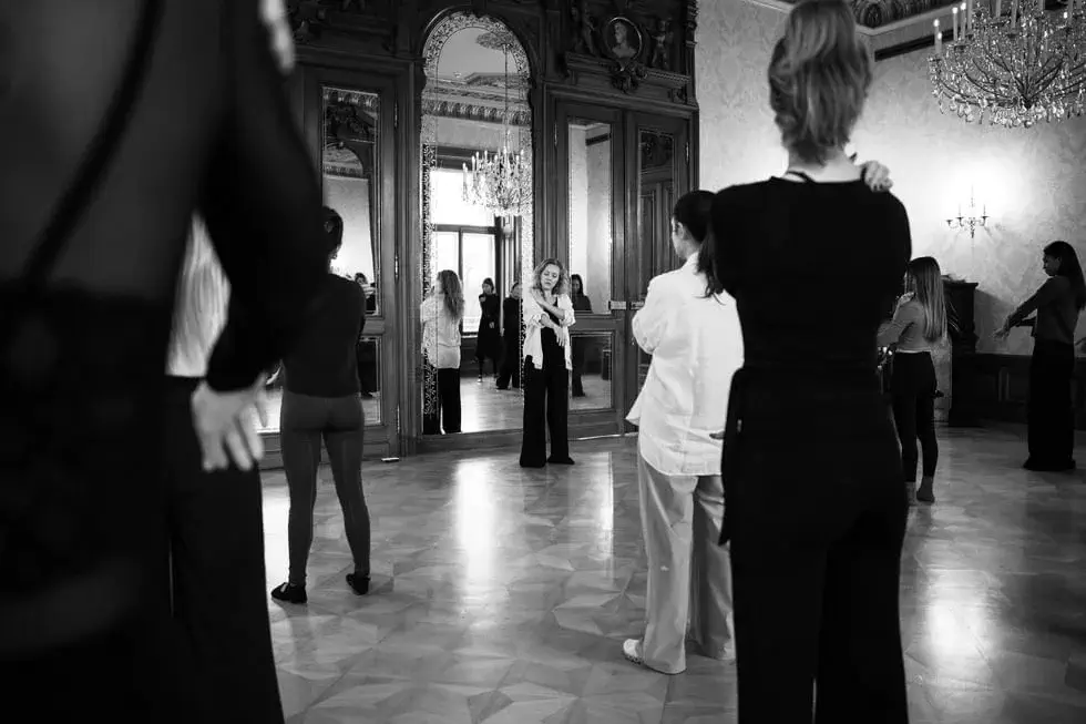 A group of people stand spaced apart in a large, ornate room with mirrors and chandeliers, facing a woman speaking at the front.
