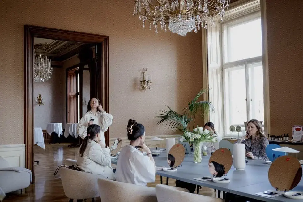 Several women are seated at a table with mirrors, makeup, and flowers in a well-lit, elegant room with chandeliers and large windows.