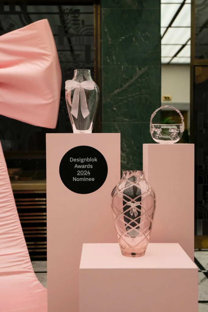 Three glass vases are displayed on pink pedestals, one with a white bow. A sign reads "Designblok Awards 2024 Nominee." Large pink fabric is draped to the left.