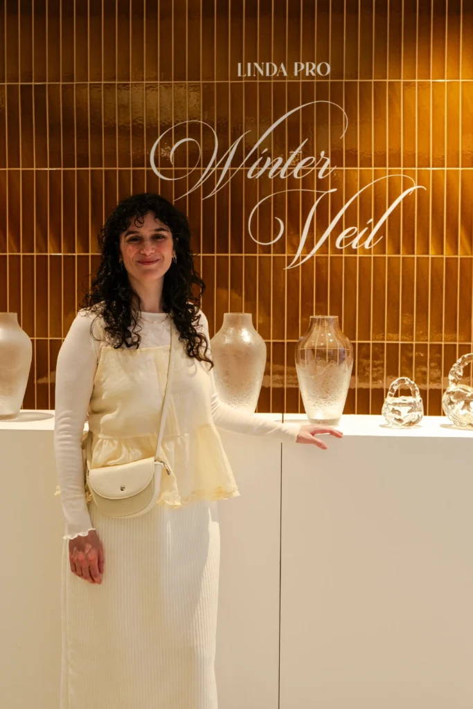 A woman stands in front of a display of glass vases, with the sign "Linda Pro Winter Veil" on a wooden wall behind her, reminiscent of an Almanac entry at Alcron Prague.