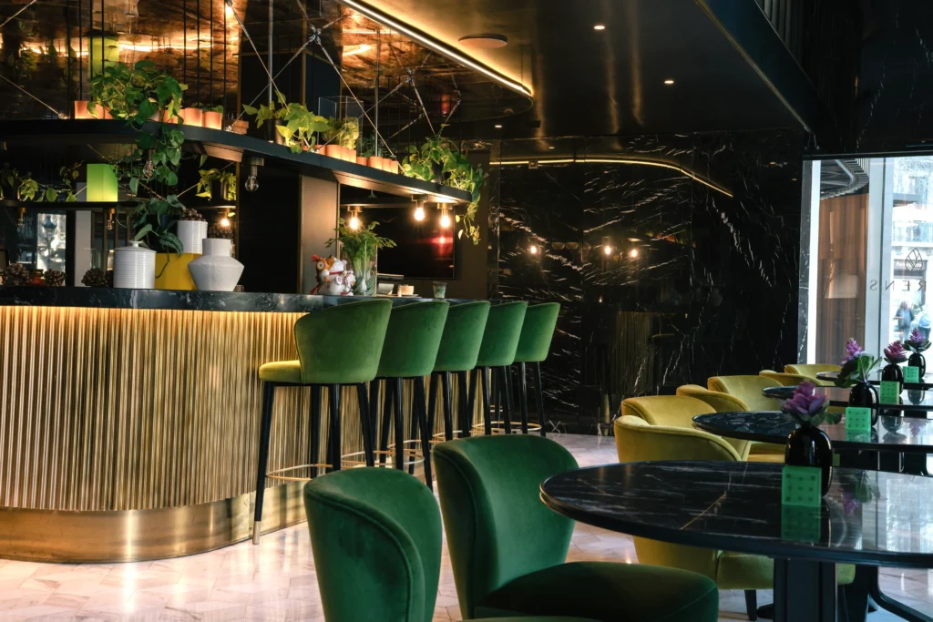 Modern bar interior with green velvet chairs, marble tables, gold accents, and hanging plants under warm lighting creates a stylish, plant forward atmosphere.