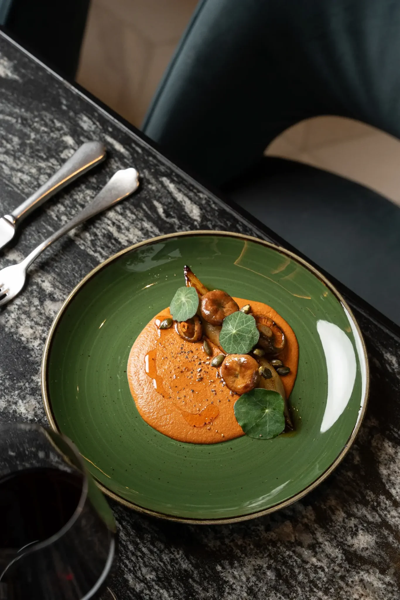 A green plate with a serving of roasted mushrooms on orange sauce, garnished with pumpkin seeds and green leaves, highlights a plant forward meal, set on a marble table beside cutlery and a glass of red wine.
