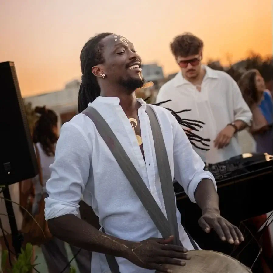At a Leo Session event, a man in a white shirt plays a drum outdoors at sunset, with a DJ and others enjoying the Zenith vibe in the background.