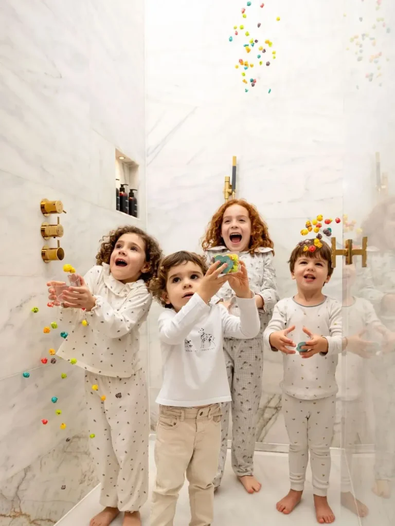 Quatre nens en pijama estan drets en una dutxa de marbre, agafant i llançant pilotes de joguina de colors o caramels que estan escampats per l'aire.