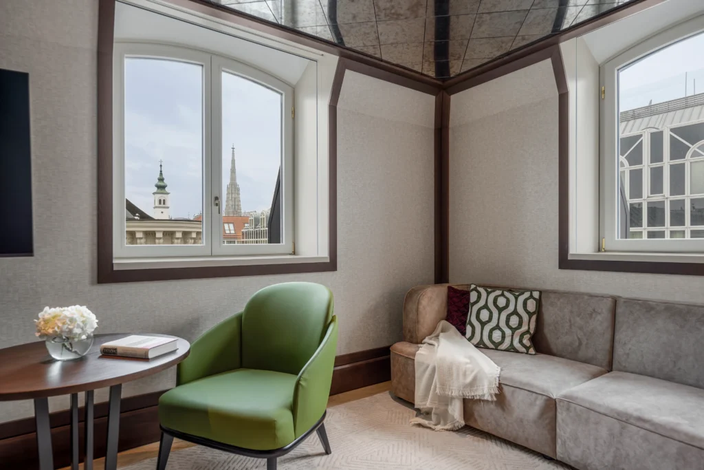 A corner of a modern room at Almanac Palais Vienna with a green chair, round table, beige sofa with cushions and throw, and two windows revealing city buildings and church spires outside.