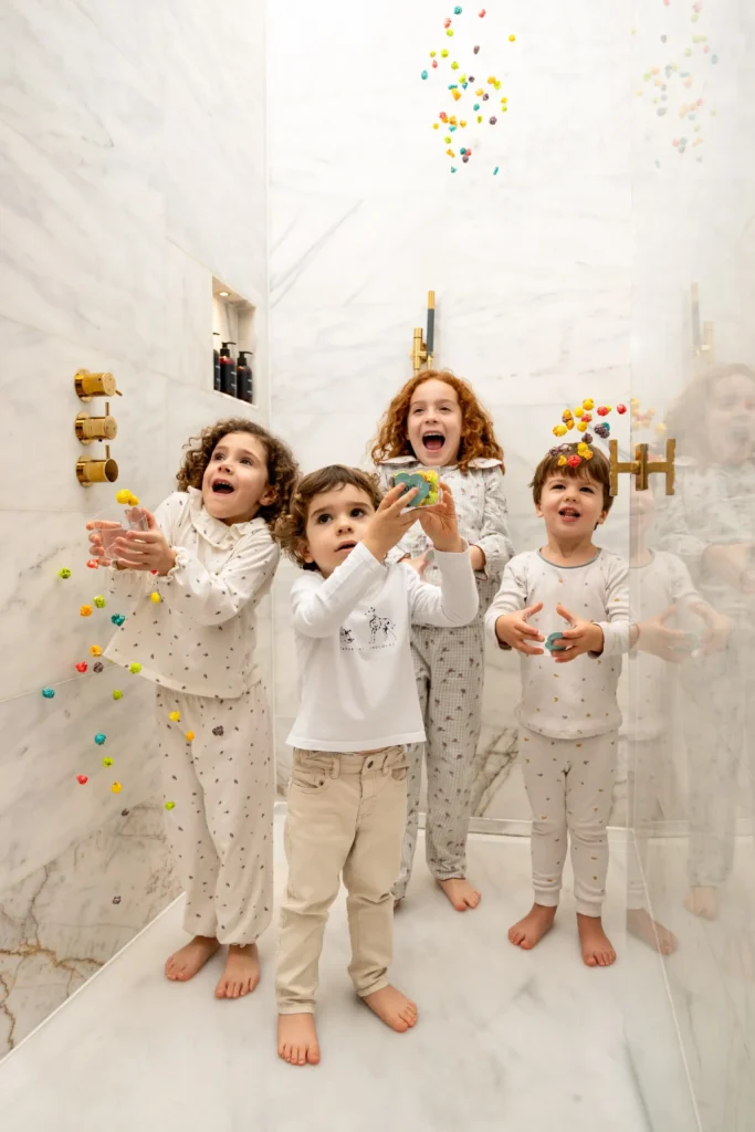 Four young children in pajamas stand in a marble shower at Almanac Barcelona, catching colorful plastic balls falling around them, with gold fixtures glinting on the wall.