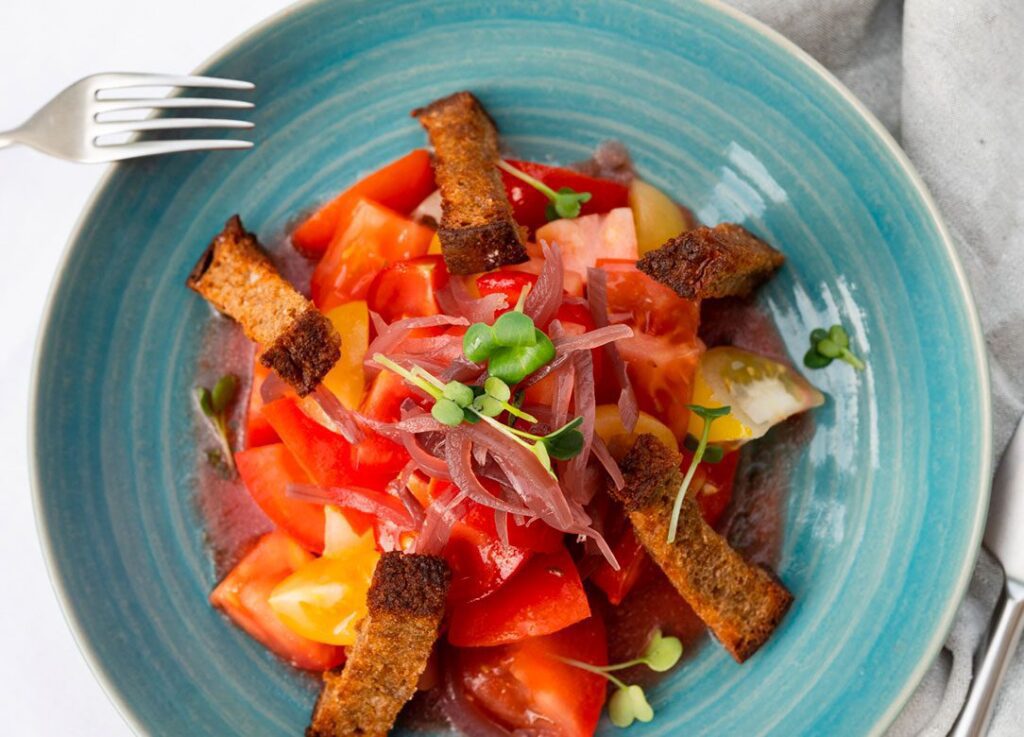 A blue plate with chopped tomatoes, pickled onions, microgreens, and toasted bread strips offers a vibrant, plant-forward meal, served with a fork and napkin beside it.