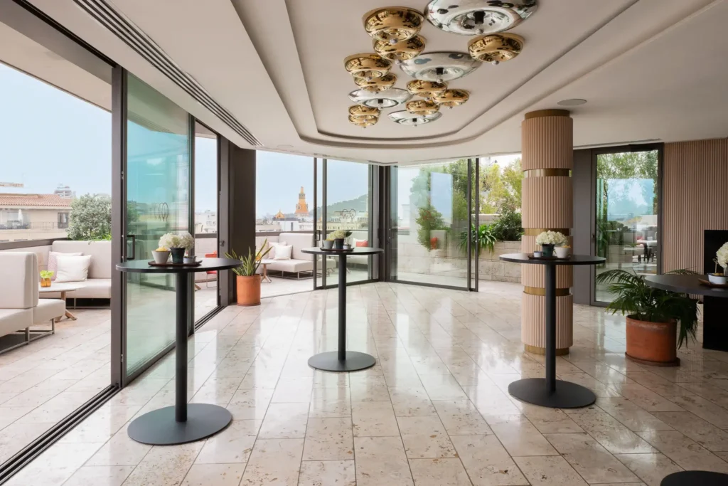 A modern indoor event space with marble floors, glass walls, tall cocktail tables, ceiling lights, and access to an outdoor terrace with seating and city views—perfect for hosting stylish events or professional meetings.