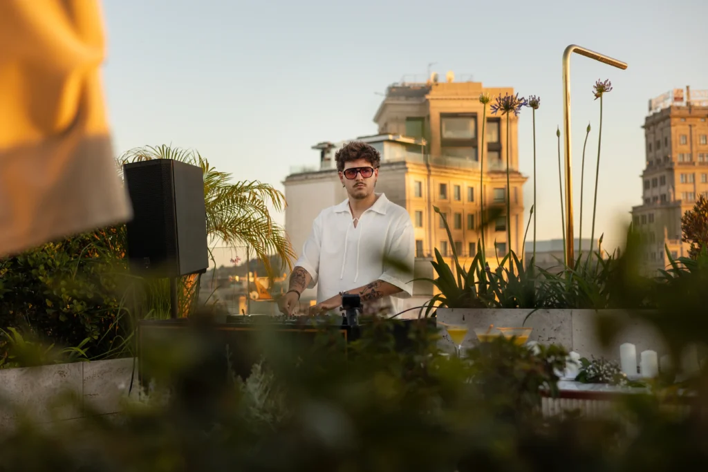 A man wearing sunglasses and a white shirt stands behind DJ equipment on a rooftop with buildings and plants in the background.