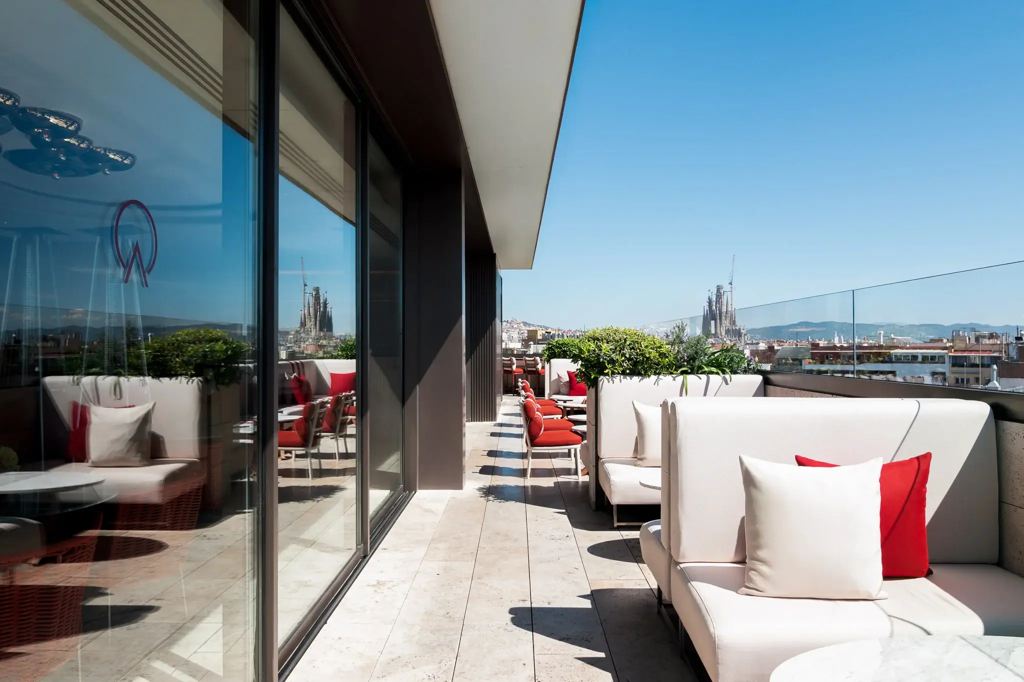 Una moderna terraza con asientos blancos acolchados, cojines rojos, plantas en macetas y una impresionante vista de la ciudad -incluida una lejana catedral- bajo un cielo azul despejado. Relájese junto a la piscina sobre la ciudad.