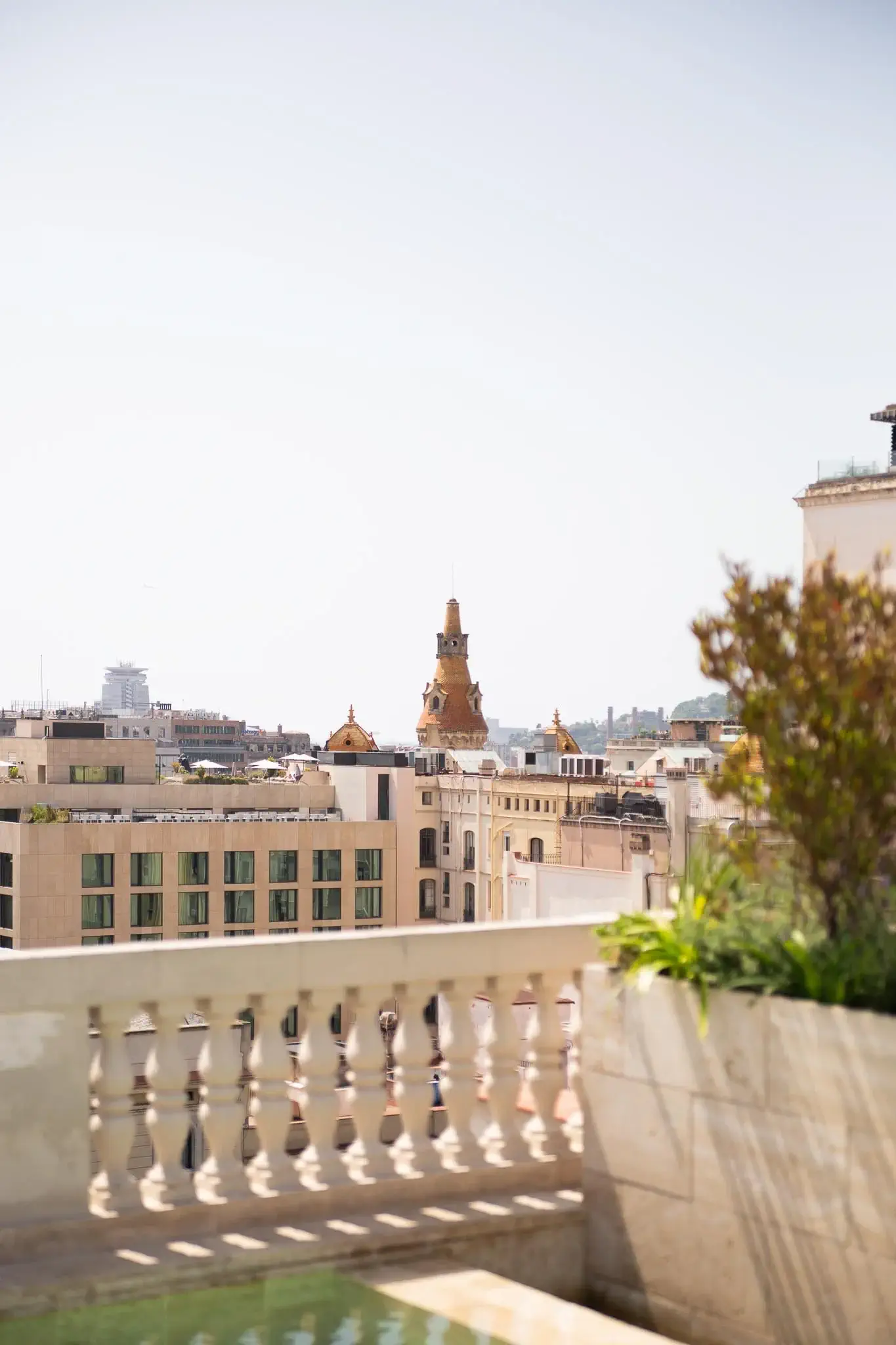Vista de un paisaje urbano con edificios de color beige, una torre decorativa al fondo y un balcón de piedra con plantas y terraza bajo un cielo despejado.