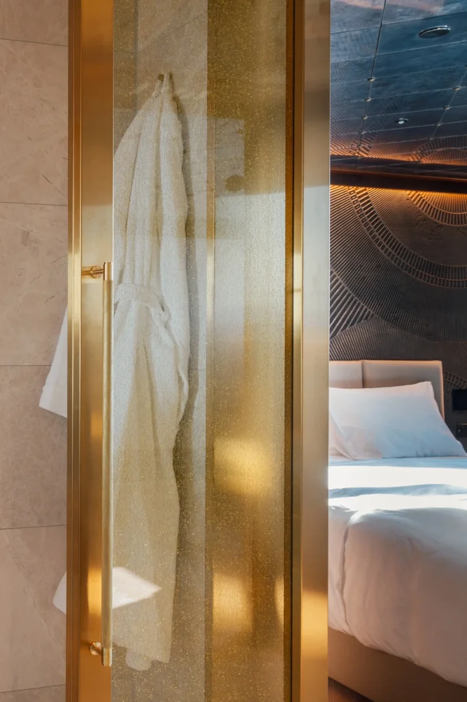 A white bathrobe hangs inside a frosted glass shower door with a gold frame, next to a bed with white linens in a modern hotel room.