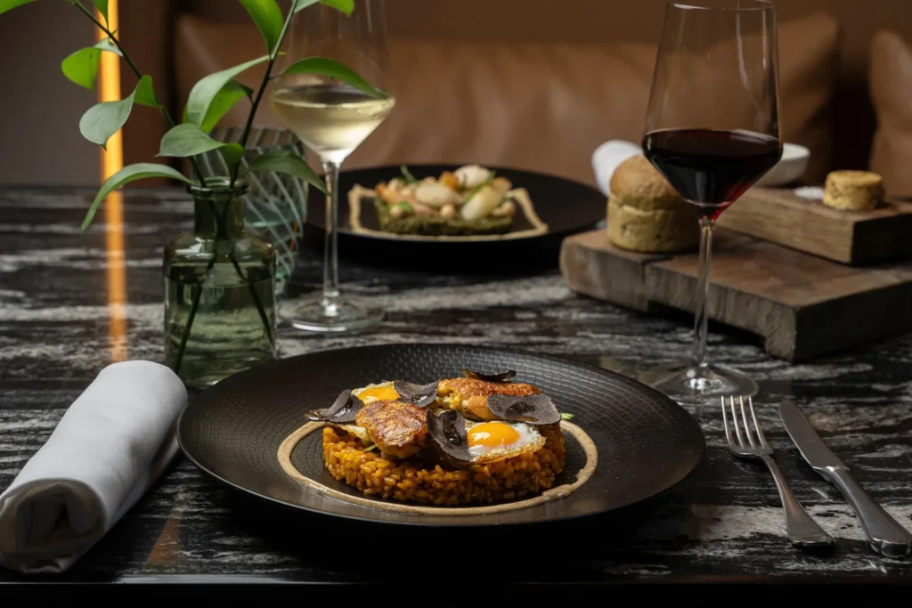 A plate of rice topped with eggs, sausage, and truffle slices sits on a marble table, accompanied by wine glasses, bread, and a side dish in the background.