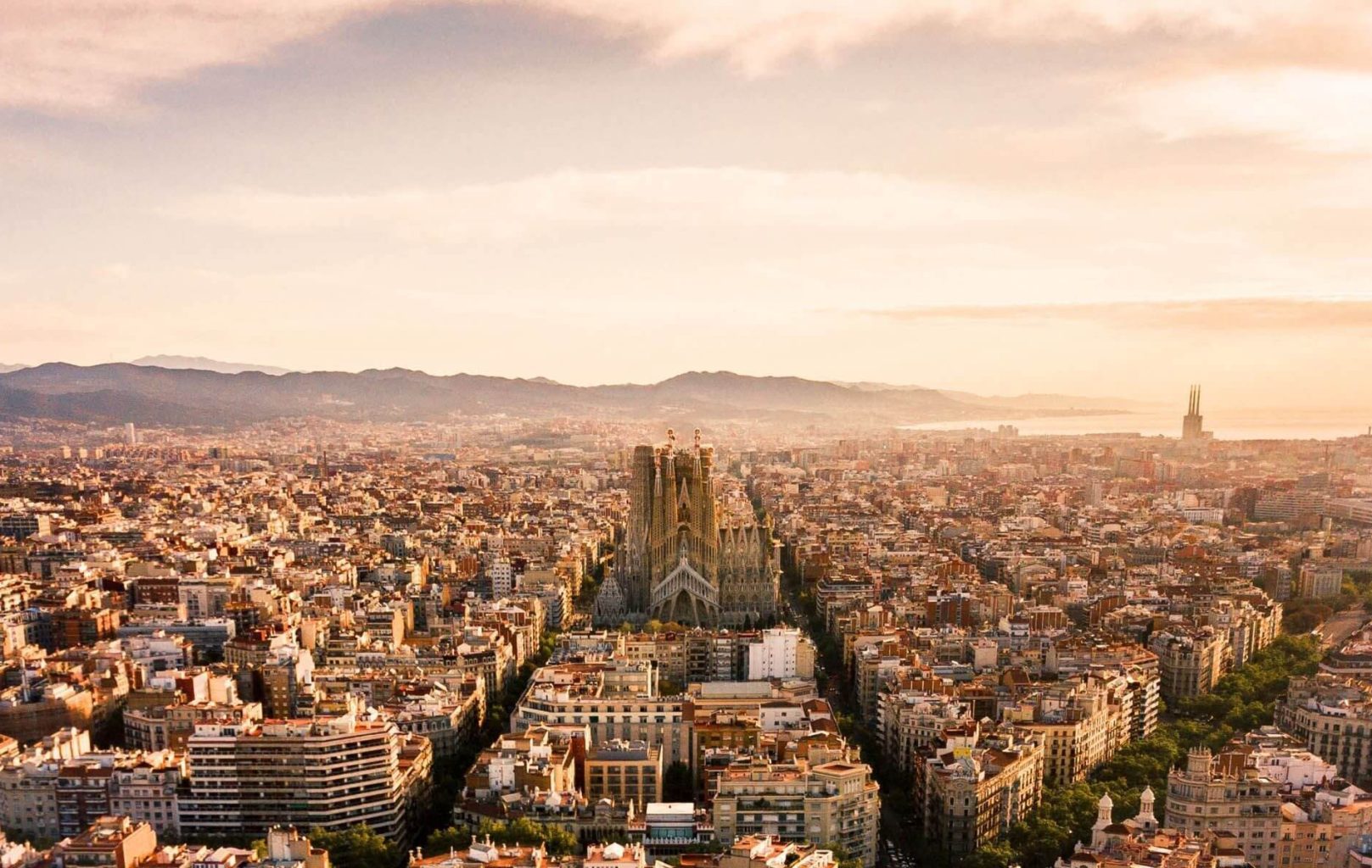 Aerial view of Barcelona at sunset, with the Sagrada Familia basilica in the center, the MNAC art museum nearby, all surrounded by city buildings and distant mountains.