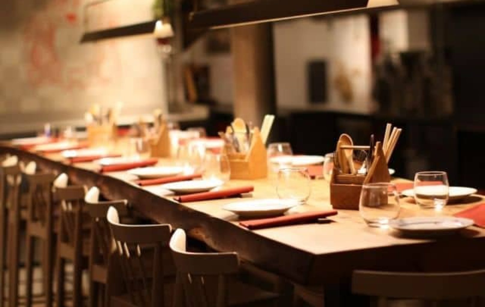Una larga mesa de madera con platos, vasos, servilletas y cubiertos en el interior de un restaurante con luz tenue.