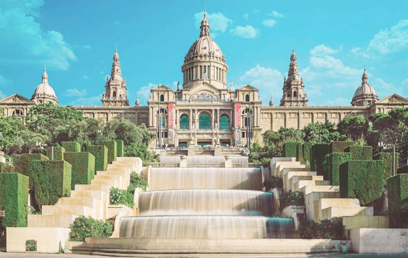 The Museu Nacional d'Art de Catalunya (MNAC) is a large historic building with a central dome and two towers, fronted by terraced gardens, staircases, and cascading fountains under a clear blue sky.