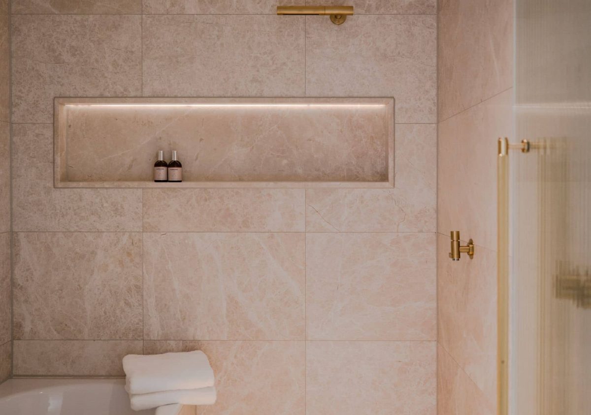 A modern bathroom in the Almanac Room features beige marble tiles, a built-in shelf with two small bottles, a folded white towel on the tub, and elegant gold fixtures.