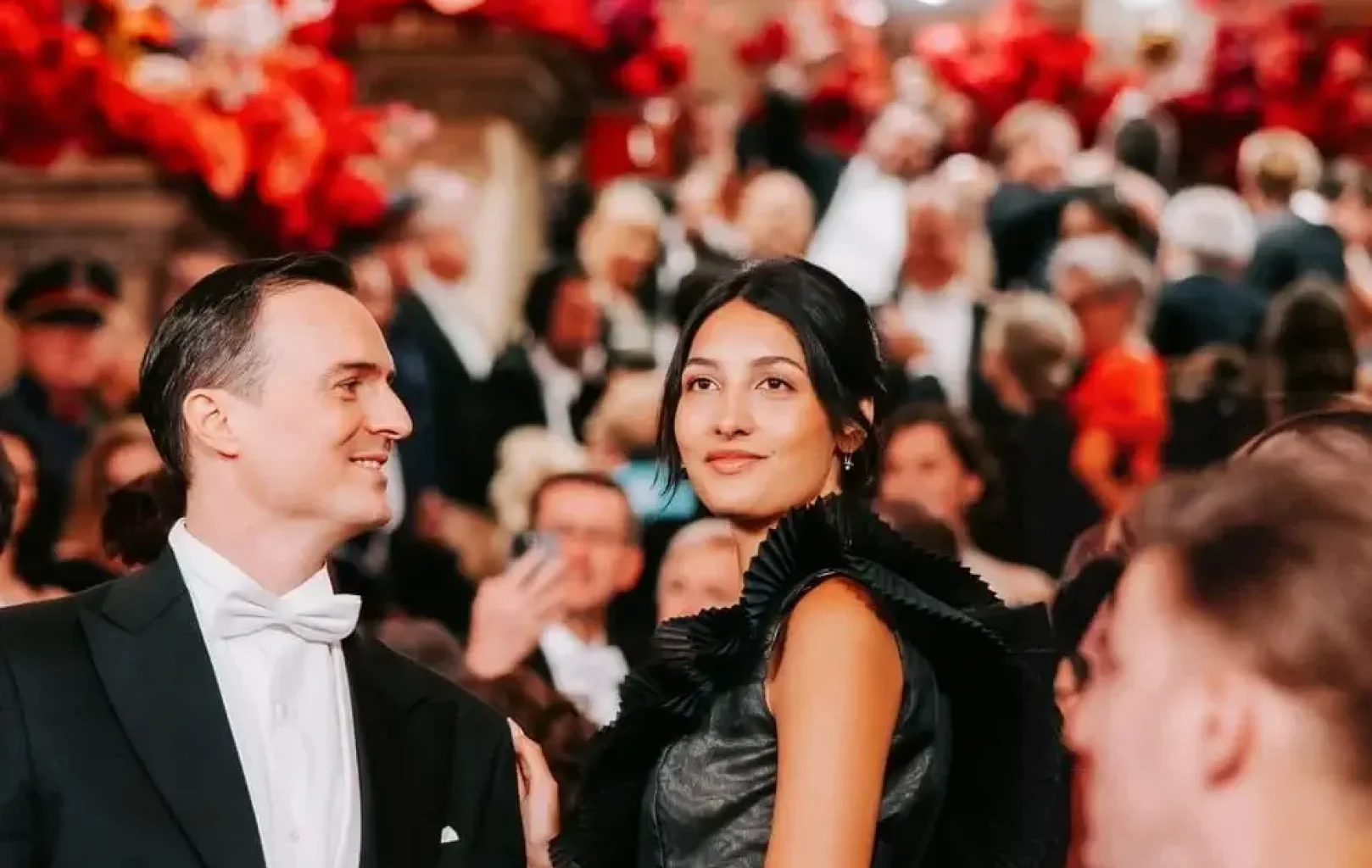 Ein gut gekleidetes Paar steht inmitten einer Menschenmenge auf einem glamourösen Nachtball, umgeben von Kronleuchtern und rotem Blumenschmuck im Hintergrund.