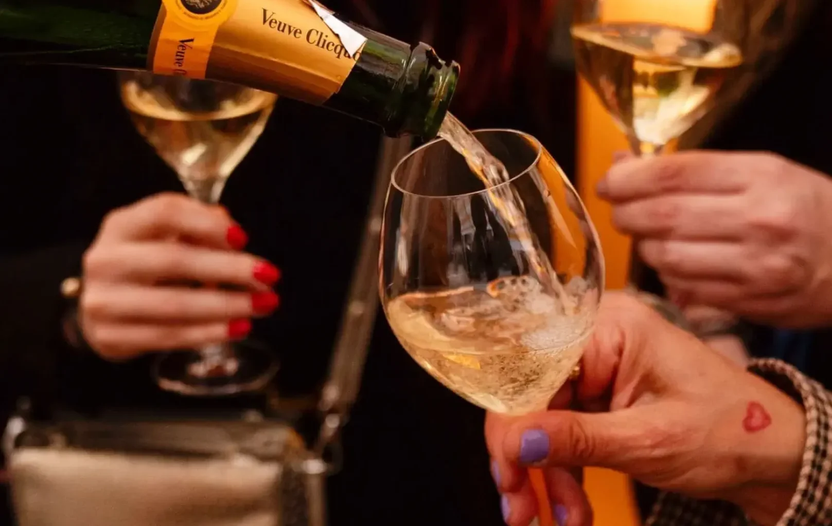 A bottle of VEUVE CLICQUOT champagne is being poured into a glass on the SUN CLUB TERRACE, while two people hold glasses of champagne in the background.