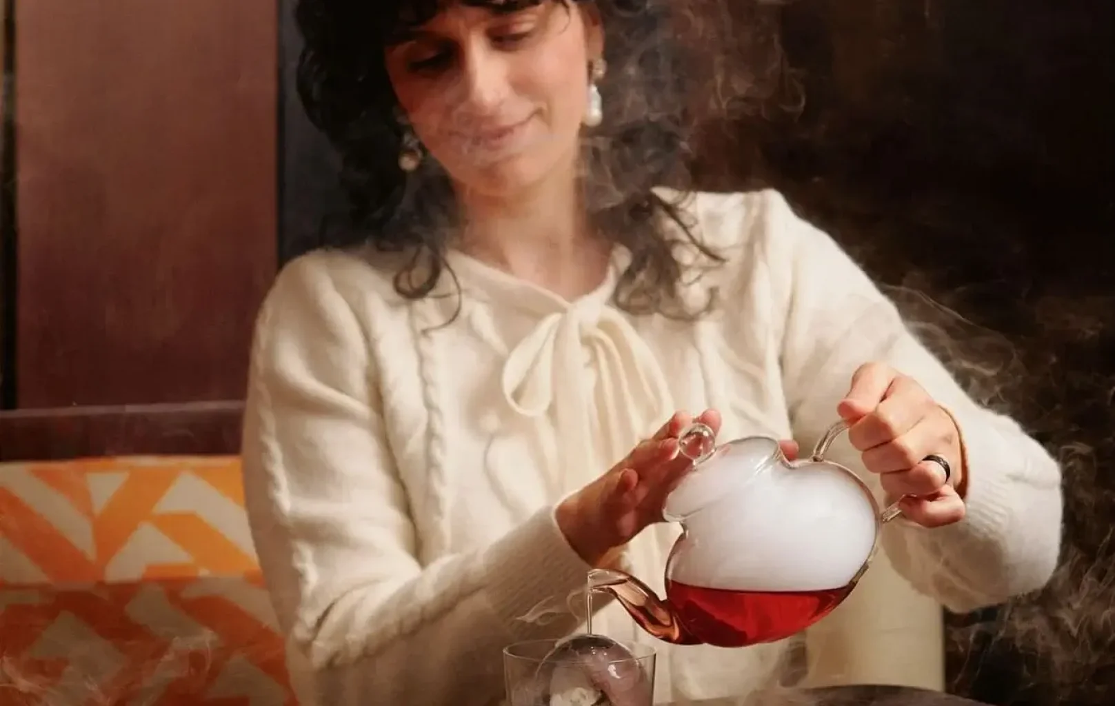 Osoba v krémovém svetru nalévá ze skleněné konvice do sklenice s velkou ledovou koulí na dřevěném stole kouřový, načervenalý koktejl SAKURA.