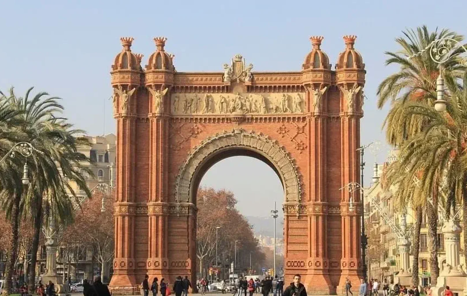 The Arc de Triomf, a red brick triumphal arch, stands in a busy public area lined with palm trees and people walking in Barcelona, especially lively in April.