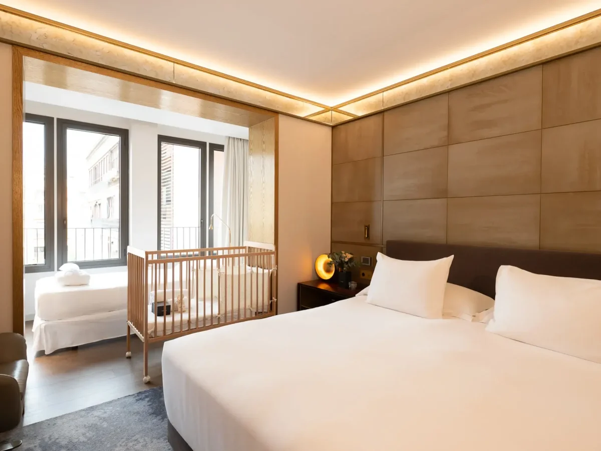 Modern accessible room with a large bed, baby crib, single bed by the window, and neutral-toned decor. Natural light floods in through floor-to-ceiling windows.