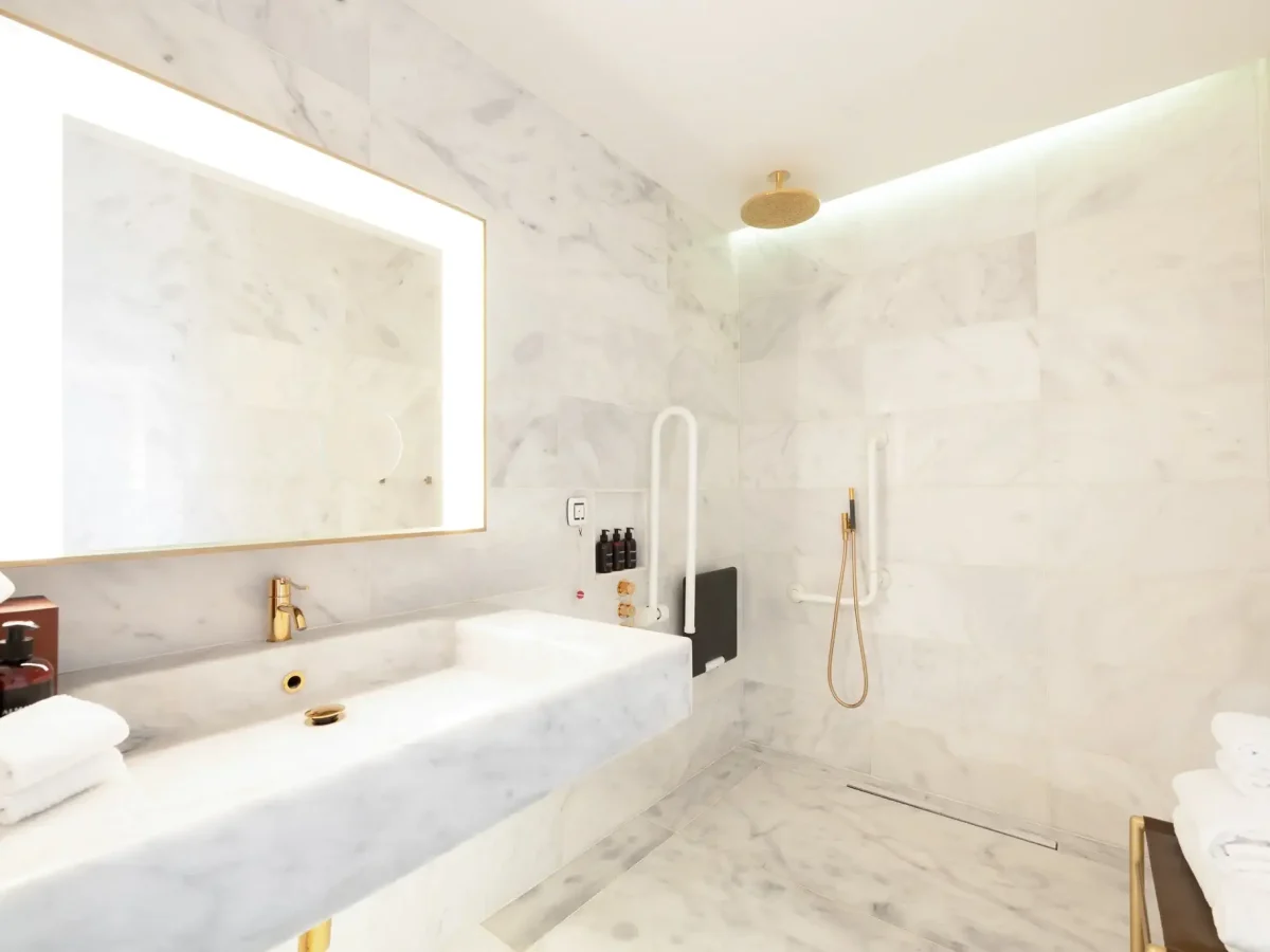 A modern Accessible Room bathroom with white marble walls, a large illuminated mirror, wide sink, walk-in shower with gold rainfall head, grab bars for safety, and neatly folded towels.