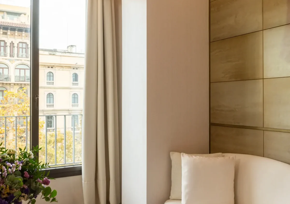 A modern Almanac Room features a white loveseat, a round wooden table with a flower vase, a large window with beige curtains leading to the balcony, and gold-toned wall panels.