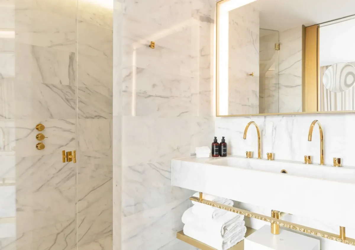 Modern bathroom with white marble walls, gold fixtures, a double sink with towels underneath, a large mirror, and a glass-enclosed shower—ideal for relaxing after enjoying the view from the balcony.