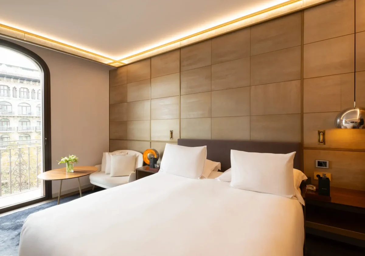 Modern Almanac Room featuring a large bed, two white chairs, a round table with flowers, and a window or balcony overlooking a building. Warm lighting and minimalist decor create an inviting atmosphere.