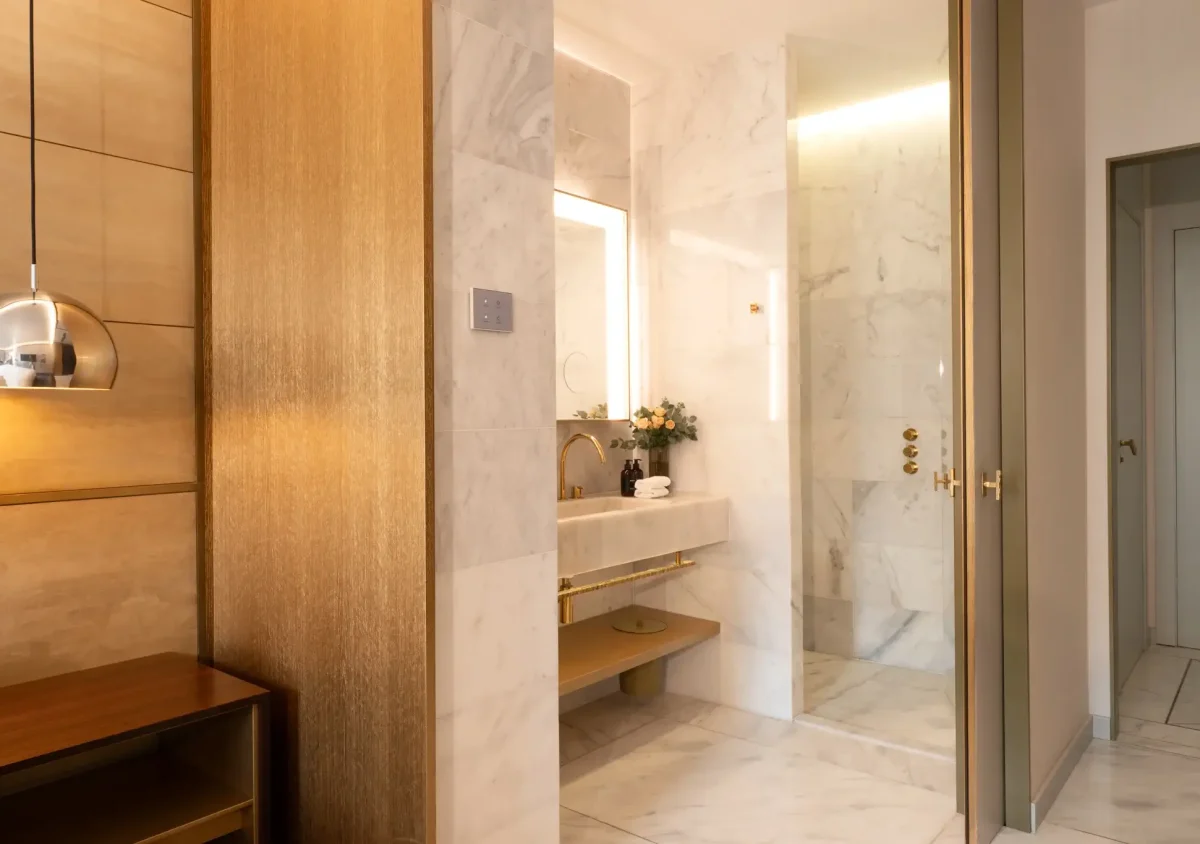 Modern bathroom with marble walls and floors, a glass shower, gold fixtures, a wooden shelving unit, and a metallic pendant light—an inviting retreat reminiscent of the luxurious style found in the Almanac Room or on an elegant balcony.