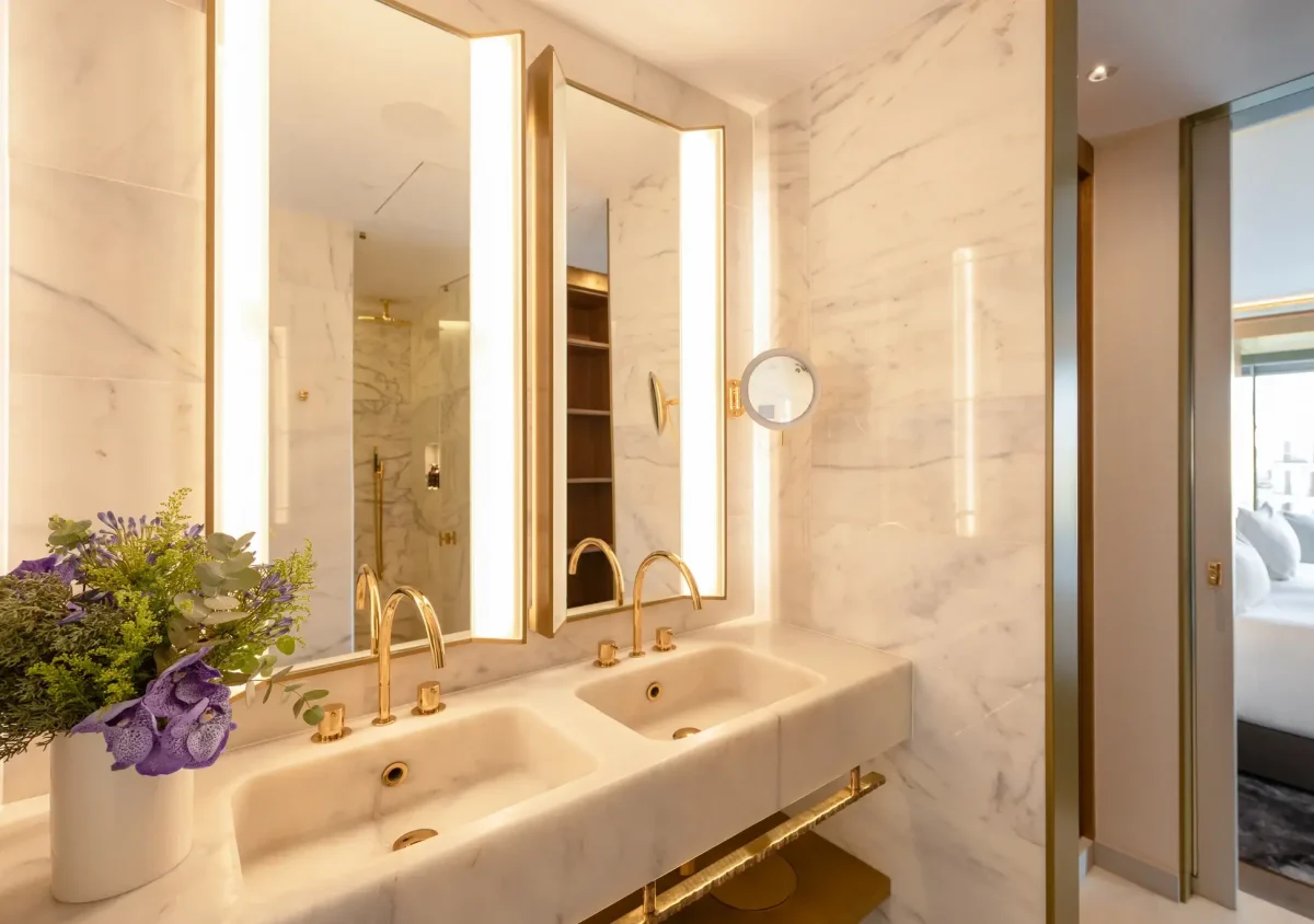 Modern bathroom with marble walls, double sink vanity with gold fixtures, large mirrors, and a bouquet of flowers on the counter; the Almanac Room bedroom is visible through the open door.