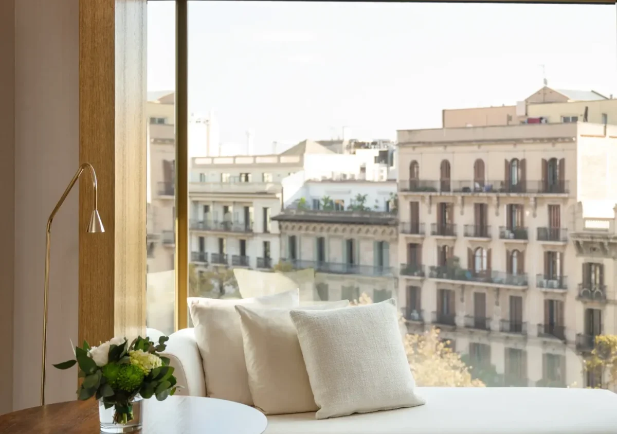 A modern Almanac Room featuring a white chaise lounge, round wooden side table, and a large window overlooking city buildings.