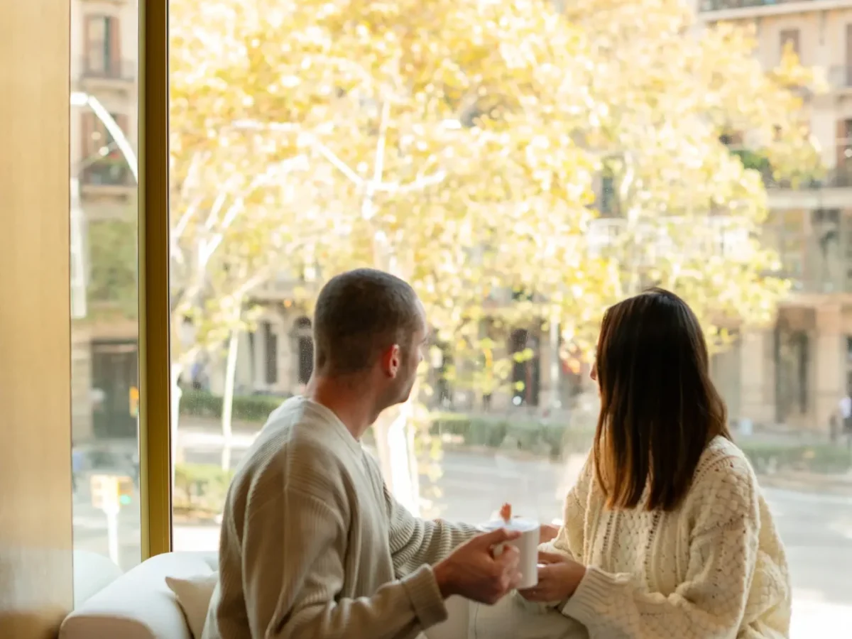 Un hombre y una mujer se sientan en un sofá blanco junto a la gran ventana de la Sala del Almanaque, sosteniendo tazas y contemplando árboles y edificios en un día soleado.