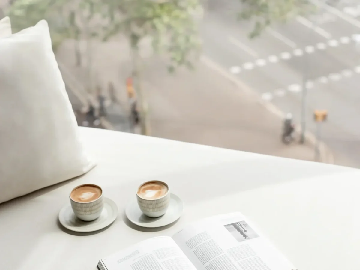 Dos tazas de café y un libro abierto descansan en el alféizar blanco de la sala del almanaque, con una calle de la ciudad visible a través de la ventana.