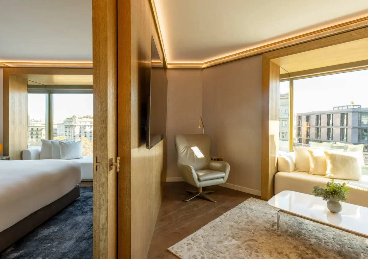 Modern Almanac Suite with a bedroom and adjoining sitting area, featuring large windows, a white armchair, a sofa, and views of city buildings outside.