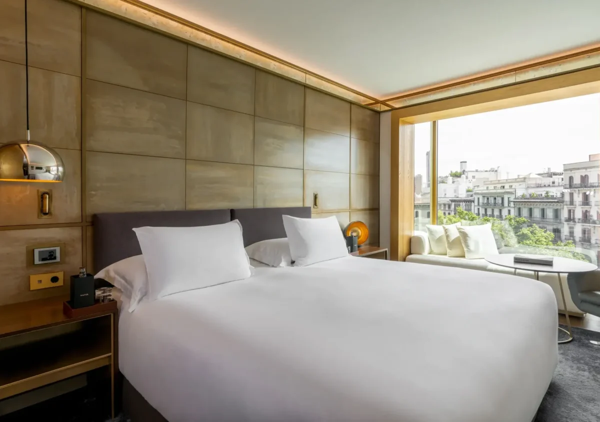 Modern Almanac Suite with a large bed, white linens, bedside tables, a hanging lamp, and a window overlooking city buildings and trees.