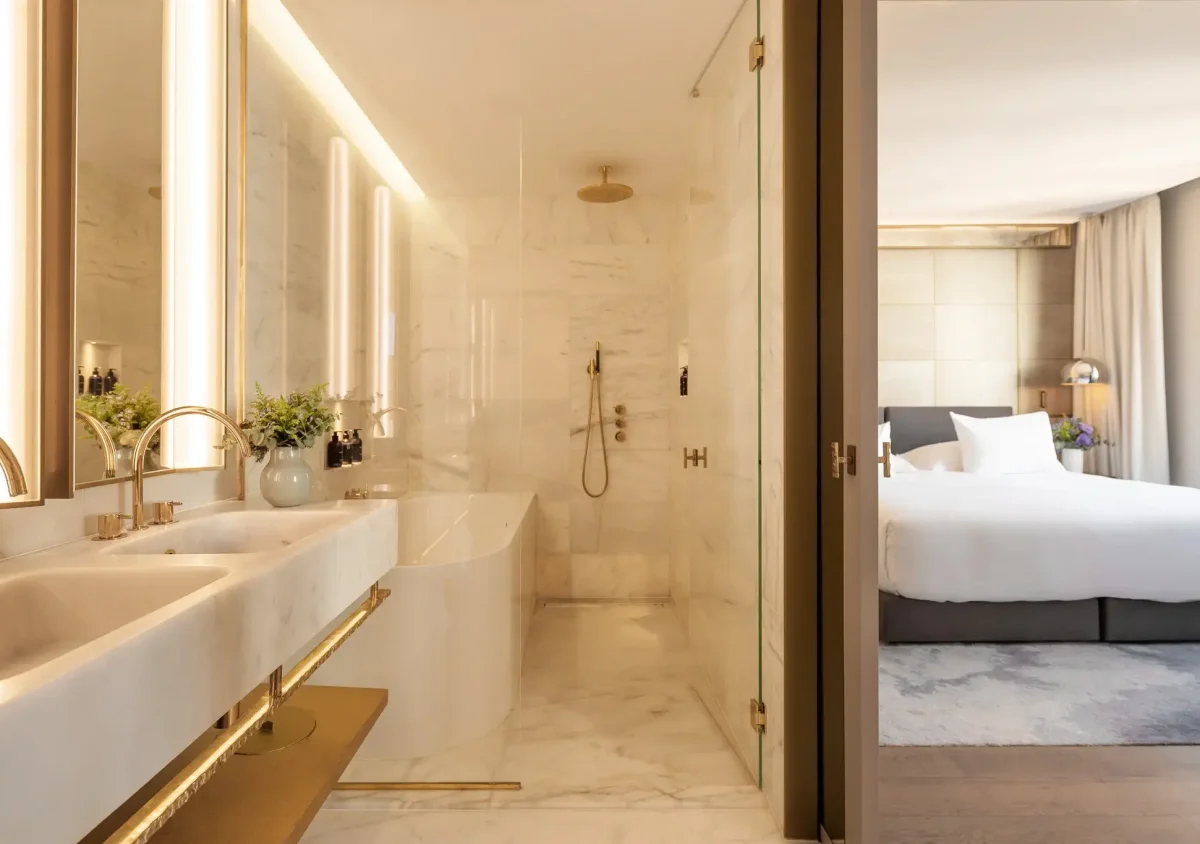 Modern Almanac Suite bathroom with double sinks, bathtub, and walk-in shower, adjacent to a bedroom featuring a large bed and abundant natural light from a window.