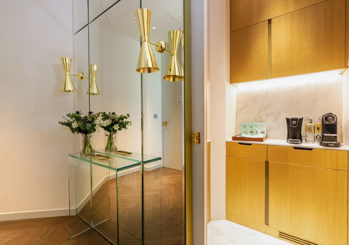 A modern entryway in the Almanac Suite features mirrored walls, gold sconces, two vases of flowers on a glass console, and an adjacent coffee station with gold cabinets and sleek coffee machines.