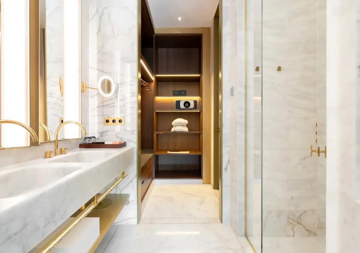 Modern Almanac Suite bathroom with marble surfaces, double sinks, gold fixtures, a glass shower, and an open closet with shelves for towels and a safe.