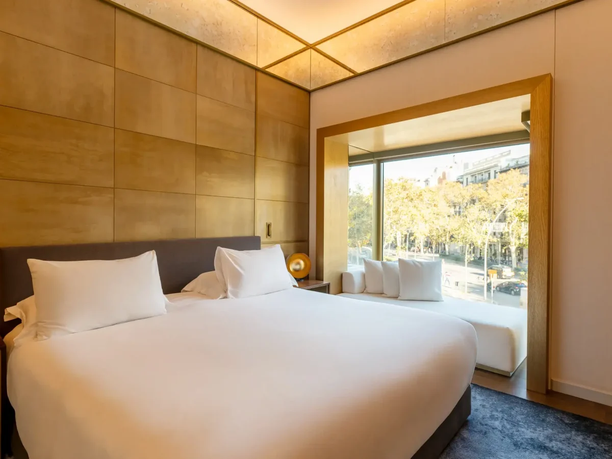 Modern Almanac Suite featuring a large bed, crisp white linens, golden wall panels, and a cozy seating area by a window overlooking a tree-lined street.