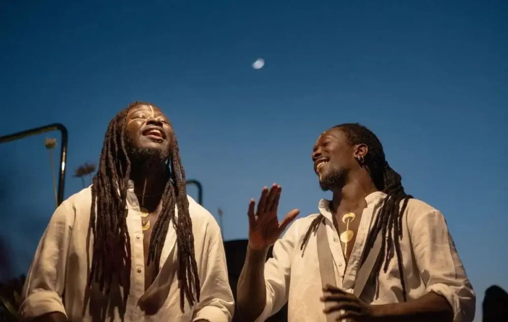 Al anochecer, dos hombres con largas rastas y camisas blancas sonríen y gesticulan con las manos. Sobre ellos se ve la luna en el cielo del atardecer.