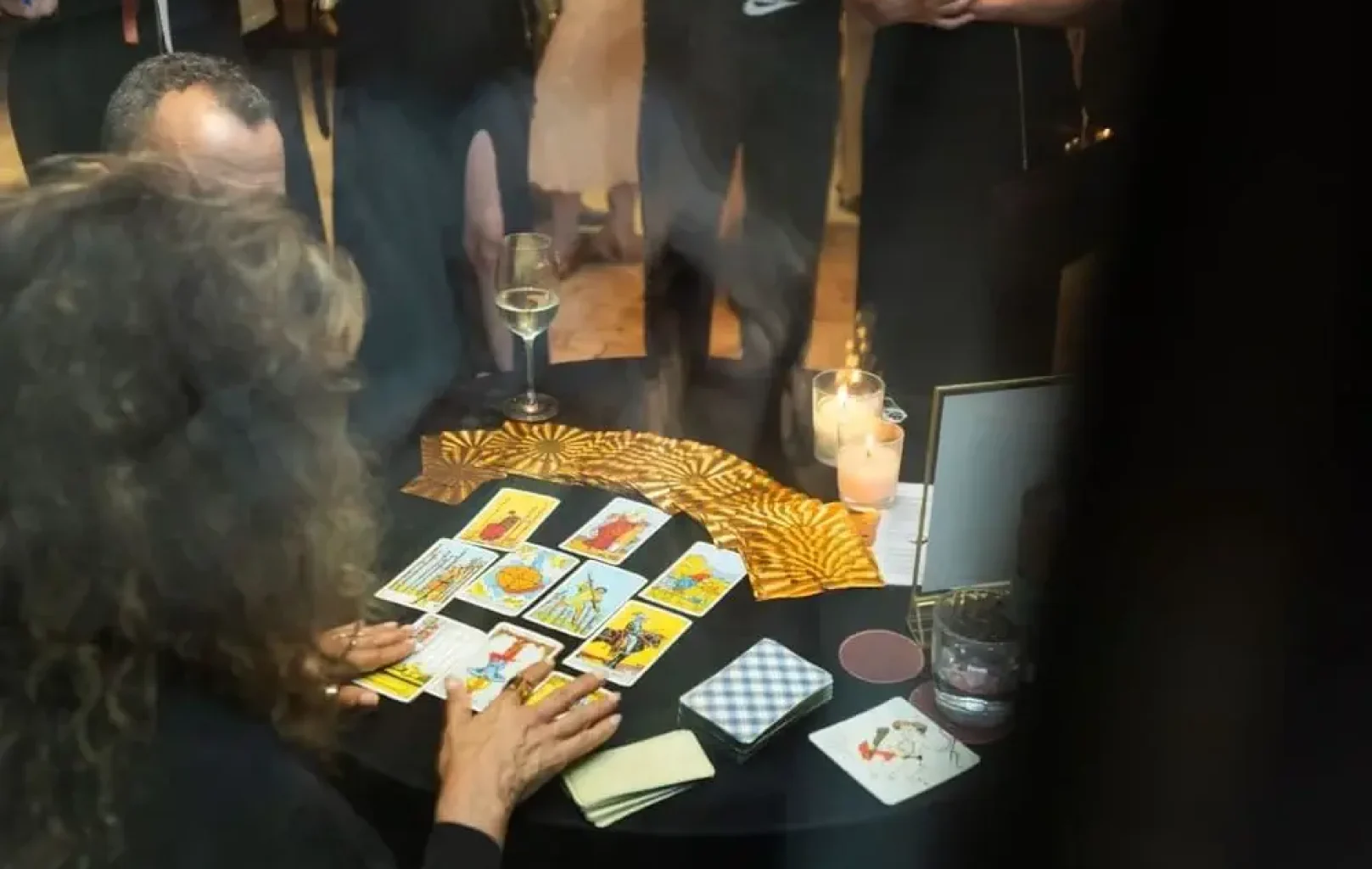 Una persona está leyendo las cartas del tarot en una mesa redonda, rodeada de gente de pie. Hay velas, bebidas y una baraja sobre la mesa.