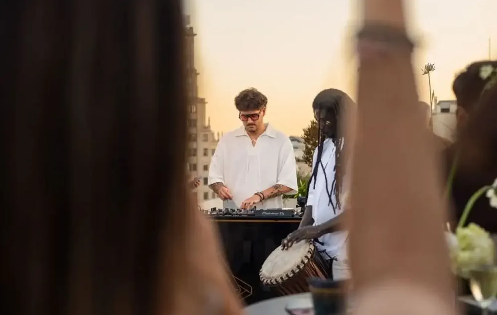 Un DJ y un percusionista actúan en un evento al aire libre en una azotea al atardecer, con miembros del público difuminados en primer plano.