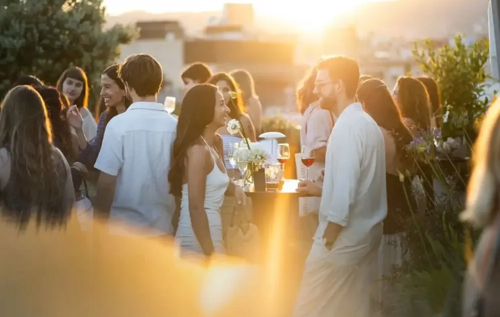 Un grupo de personas se reúne al aire libre al atardecer, charlando y tomando copas alrededor de una mesa decorada con flores.