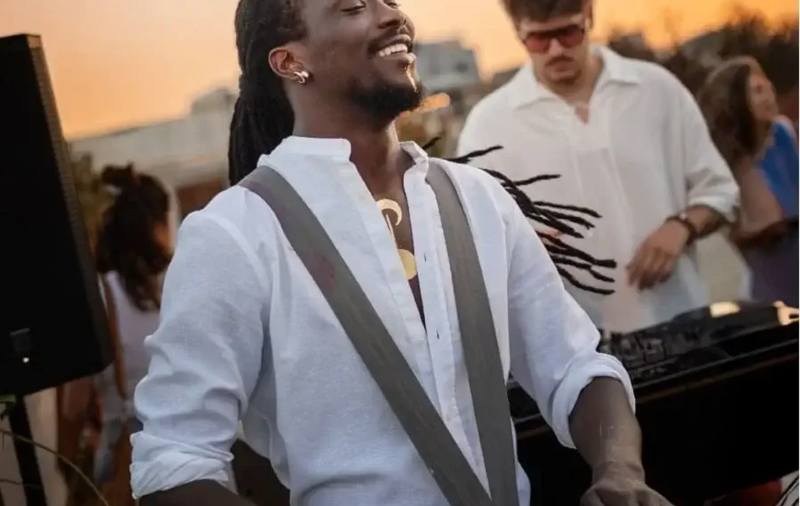 Un hombre con camisa blanca toca un tambor al aire libre al atardecer, sonriendo, con gente y un DJ de fondo.